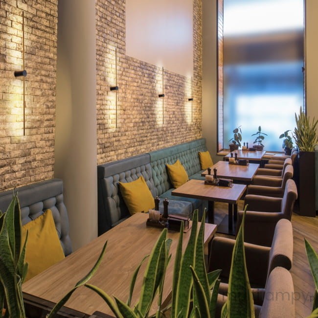 Kinkiet Wand 467 z dwoma lampami LED 10W w kolorze czarnym i złotym. Zamontowany na ścianie w restauracji, oświetla przestrzeń nad siedzeniami z niebieską tapicerką i żółtymi poduszkami, w otoczeniu roślin doniczkowych.