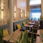 Kinkiet Wand 467 z dwoma lampami LED 10W w kolorze czarnym i złotym. Zamontowany na ścianie w restauracji, oświetla przestrzeń nad siedzeniami z niebieską tapicerką i żółtymi poduszkami, w otoczeniu roślin doniczkowych.
