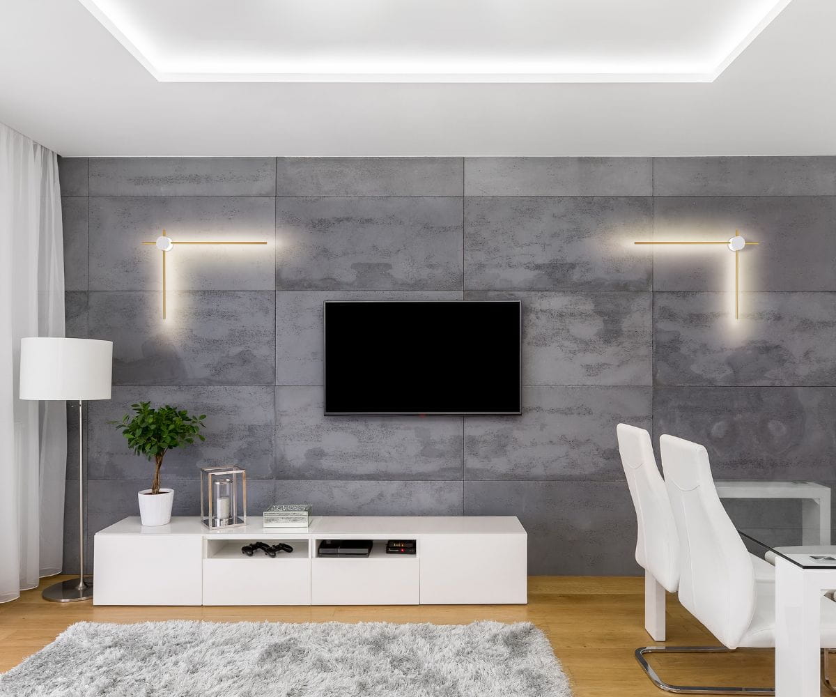Kinkiet Wand 469 z dwoma źródłami LED 12W w kolorze białym i złotym, zamontowany na szarej ścianie w nowoczesnym wnętrzu. Prosta, geometryczna forma z okrągłym elementem na końcu. W tle telewizor i biały zestaw meblowy.