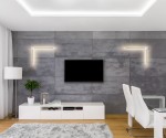 Kinkiet Wand 469 z dwoma źródłami LED 12W w kolorze białym i złotym, zamontowany na szarej ścianie w nowoczesnym wnętrzu. Prosta, geometryczna forma z okrągłym elementem na końcu. W tle telewizor i biały zestaw meblowy.