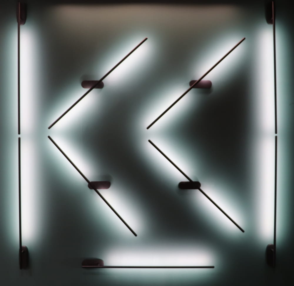 Kinkiet LED Line 243A o mocy 10W w kolorze czarnym. Produkt montowany na ścianie, składający się z ukośnych linii świetlnych, emitujących delikatne światło. Widoczne mocowania w formie prostokątnych uchwytów. Tło jasne, co podkreśla efekt świetlny.