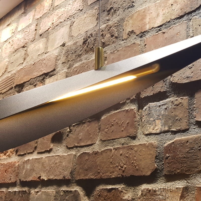 Lampa wisząca LED Buket 357, czarna z złotym akcentem, w kształcie płaskiego, lekko zakrzywionego panelu. Zawieszona na cienkim przewodzie, emituje ciepłe światło. Tło stanowi surowa, ceglasta ściana, podkreślająca nowoczesny design.