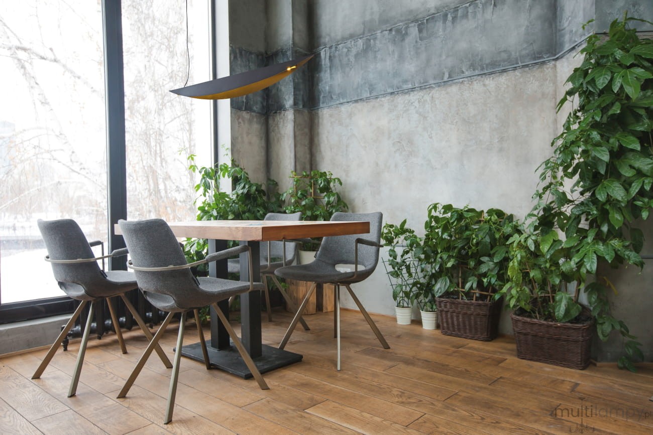 Lampa wisząca LED Buket 357 w kolorze czarnym i złotym, zawieszona w pomieszczeniu, nad drewnianym stołem. Widoczne metalowe detale i geometryczny kształt. W tle roślinność w doniczkach oraz nowoczesne krzesła.