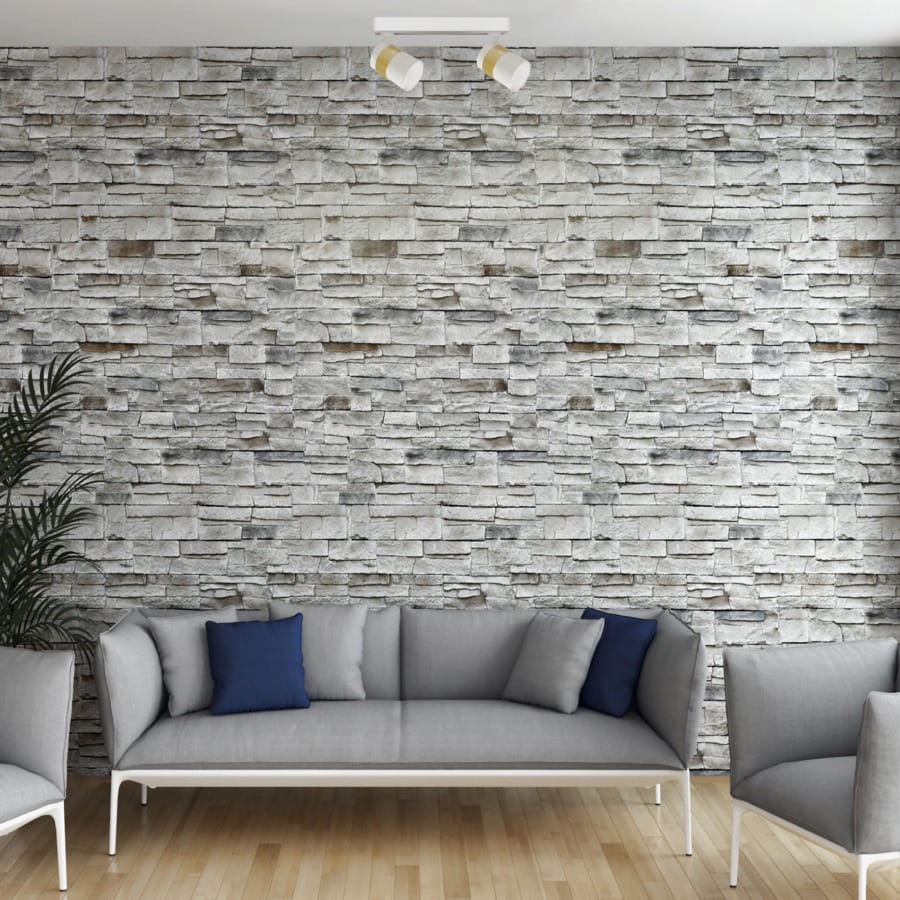 Lampa sufitowa Anillo (spot) 2xGU10 w kolorze białym, zamontowana na suficie, z dwoma regulowanymi źródłami światła. Tło stanowi ściana z kamienia, a przedmiot znajduje się nad szarym sofą z niebieskimi poduszkami.