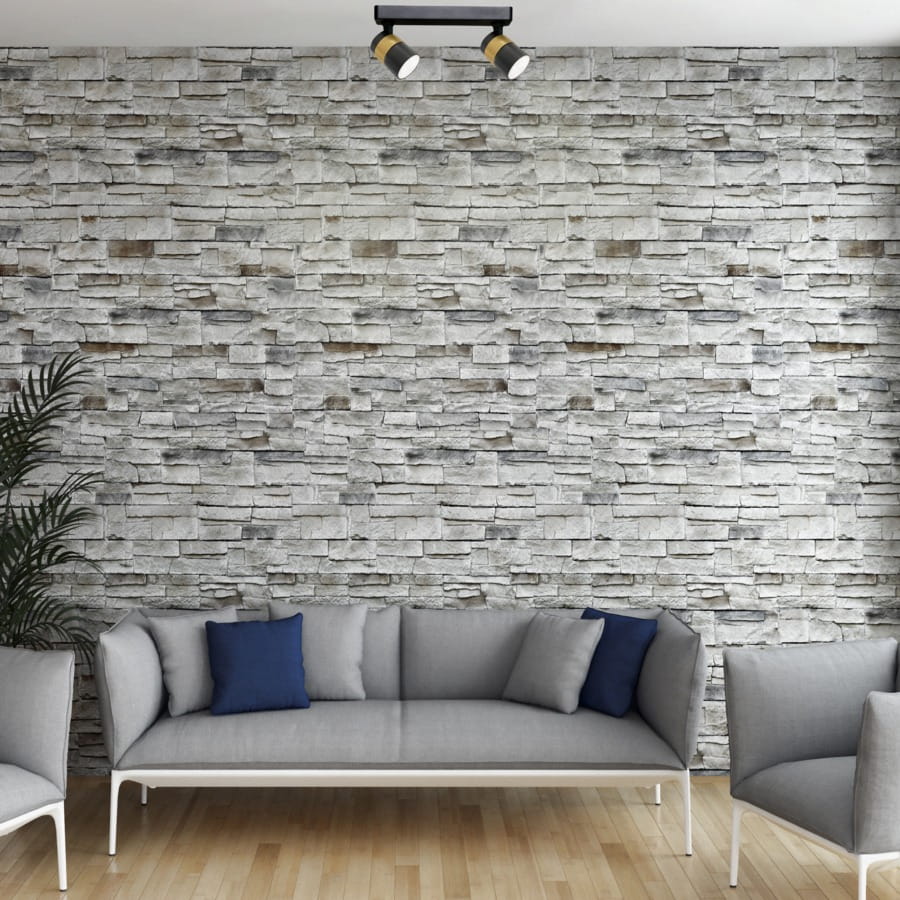 Lampa sufitowa Anillo, czarna, z dwoma spotami GU10. Ujęcie z góry na zamontowaną lampę na tle kamiennej ściany. W tle widoczna szara sofa z niebieskimi poduszkami oraz roślina.