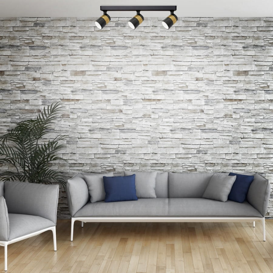 Lampa sufitowa Anillo (spot) 3xGU10 w kolorze czarnym, zamontowana na suficie, z trzema reflektorami kierunkowymi. Tło stanowi kamienna ściana, a w aranżacji widoczne są szare meble z niebieskimi poduszkami.
