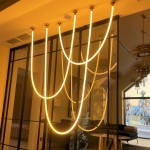 Lampa wisząca Rope Light 1xLED/73W w złotym kolorze, z kilkoma wstęgami światła w kształcie łuków, zamontowana na suficie. W tle widoczna szklana ściana i elementy wystroju wnętrza, co podkreśla nowoczesny styl aranżacji.