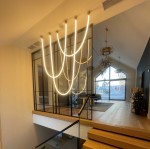 Lampa wisząca Rope Light z jednym źródłem światła LED o mocy 73W w złotym kolorze. Ułożona w kształcie fal, zawieszona na czterech punktach, w nowoczesnym wnętrzu z drewnianą podłogą i szklanymi elementami.