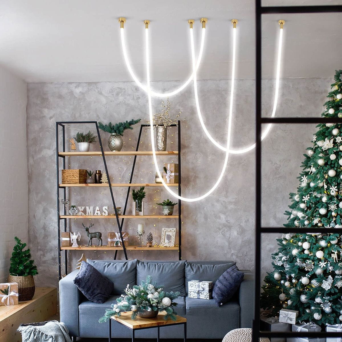 Lampa wisząca Rope Light 1xLED/50W w kolorze złotym, z dwoma smukłymi, białymi przewodami świetlnymi, zawieszona w nowoczesnym wnętrzu z szaro-beżowymi ścianami, w pobliżu dekoracji świątecznych i sofy.