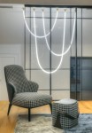Lampa wisząca Rope Light 1xLED/50W w złotym wykończeniu, zawieszona w formie łuków, z oświetleniem LED w kształcie wstęgi. W tle czarna, szklana ścianka oraz tapicerowane krzesło w wzór pied de poule na drewnianej podłodze.