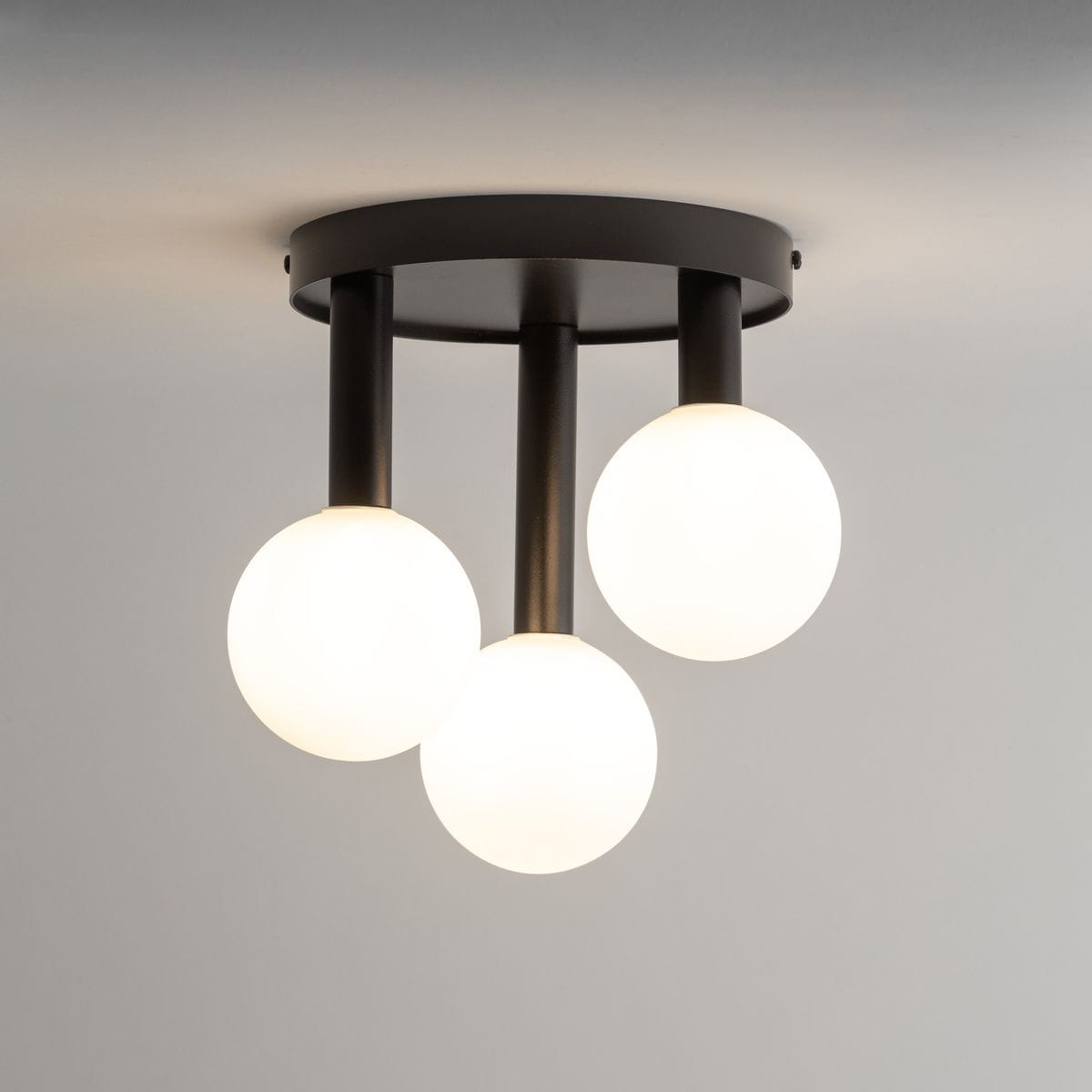 Czarna lampa sufitowa Perla z trzema białymi kulistymi kloszami na rurkach, zamocowana na okrągłej podstawie. Klosze wykonane z matowego szkła, emitujące ciepłe światło. Ujęcie z dołu, zbliżenie na detal konstrukcji.