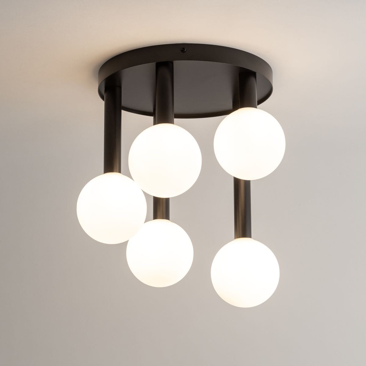 Lampa sufitowa Perla z pięcioma źródłami światła G9, czarna z białymi kulistymi kloszami. Ułożona w poziomie, z prostymi, metalowymi ramionami. Tło jasne, jednolite. Wymiary i szczegóły montażowe nieokreślone.