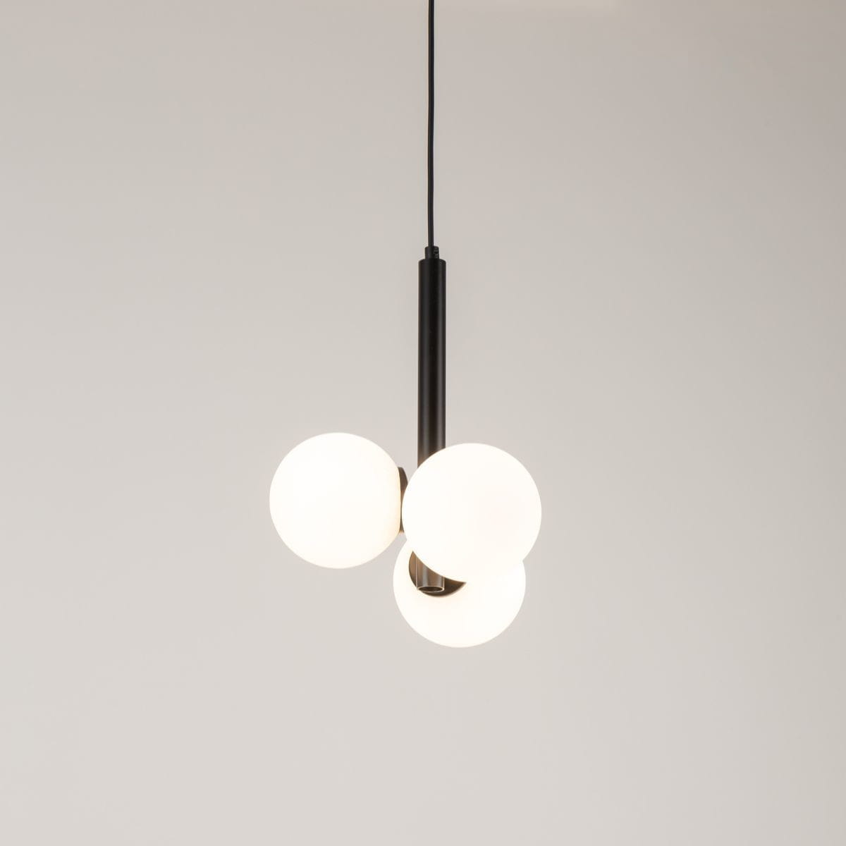 Lampa wisząca Perla z trzema kulistymi kloszami z białego szkła, zawieszona na czarnym metalowym stelażu. Klosze w układzie trójwymiarowym, wiszące na cienkim przewodzie. Tło jasne, minimalistyczne.