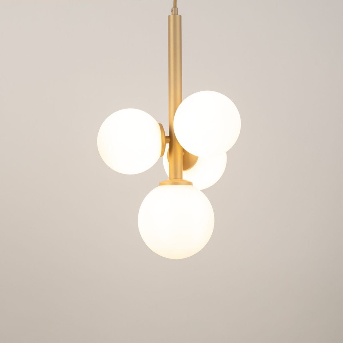 Lampa wisząca Perla z czterema okrągłymi, mlecznymi kloszami na złotej konstrukcji. Ujęcie z góry, prezentujące elegancki design. Klosze rozmieszczone asymetrycznie, zawieszone na długim, prostym ramieniu. Tło w jasnym kolorze.