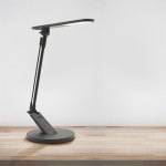 Czarna lampka biurkowa Optimum LED 7W/400lm z regulowanym ramieniem i okrągłą podstawą. Widoczny panel sterujący na podstawie. Ujęcie z boku, na jasnym tle drewnianego stołu.