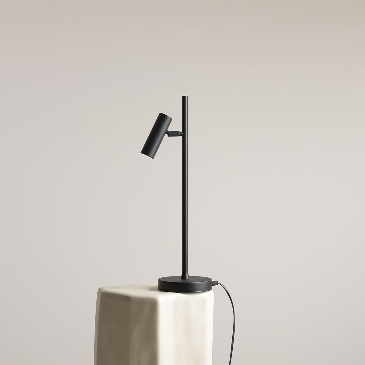Lampka biurkowa Trevo w kolorze czarnym, o minimalistycznym designie. Posiada cienką, prostą nogę oraz regulowany klosz w formie cylinderka. Ustawiona na jasnej, geometrycznej podstawie. Widoczny kabel zasilający. Tło w neutralnym odcieniu.