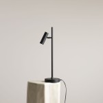 Lampka biurkowa Trevo w kolorze czarnym, o minimalistycznym designie. Posiada cienką, prostą nogę oraz regulowany klosz w formie cylinderka. Ustawiona na jasnej, geometrycznej podstawie. Widoczny kabel zasilający. Tło w neutralnym odcieniu.