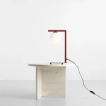 Czerwona lampa stołowa Zac z jedną oprawą, z metalowym stelażem w kształcie litery L oraz okrągłą, matową kloszem. Lampa stoi na prostokątnej, jasnej podstawie z kamienia. Widoczny czarny kabel z włącznikiem. Tło jasne, minimalistyczne.
