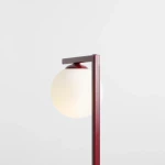 Czerwona lampa stołowa Zac 1-punktowa z białym, okrągłym kloszem. Ujęcie z boku, pokazujące prostą, metalową konstrukcję z poziomą poprzeczką. Klosz umieszczony na górze, lekko przemawia przez matowe szkło. Tło jednolite, jasne.