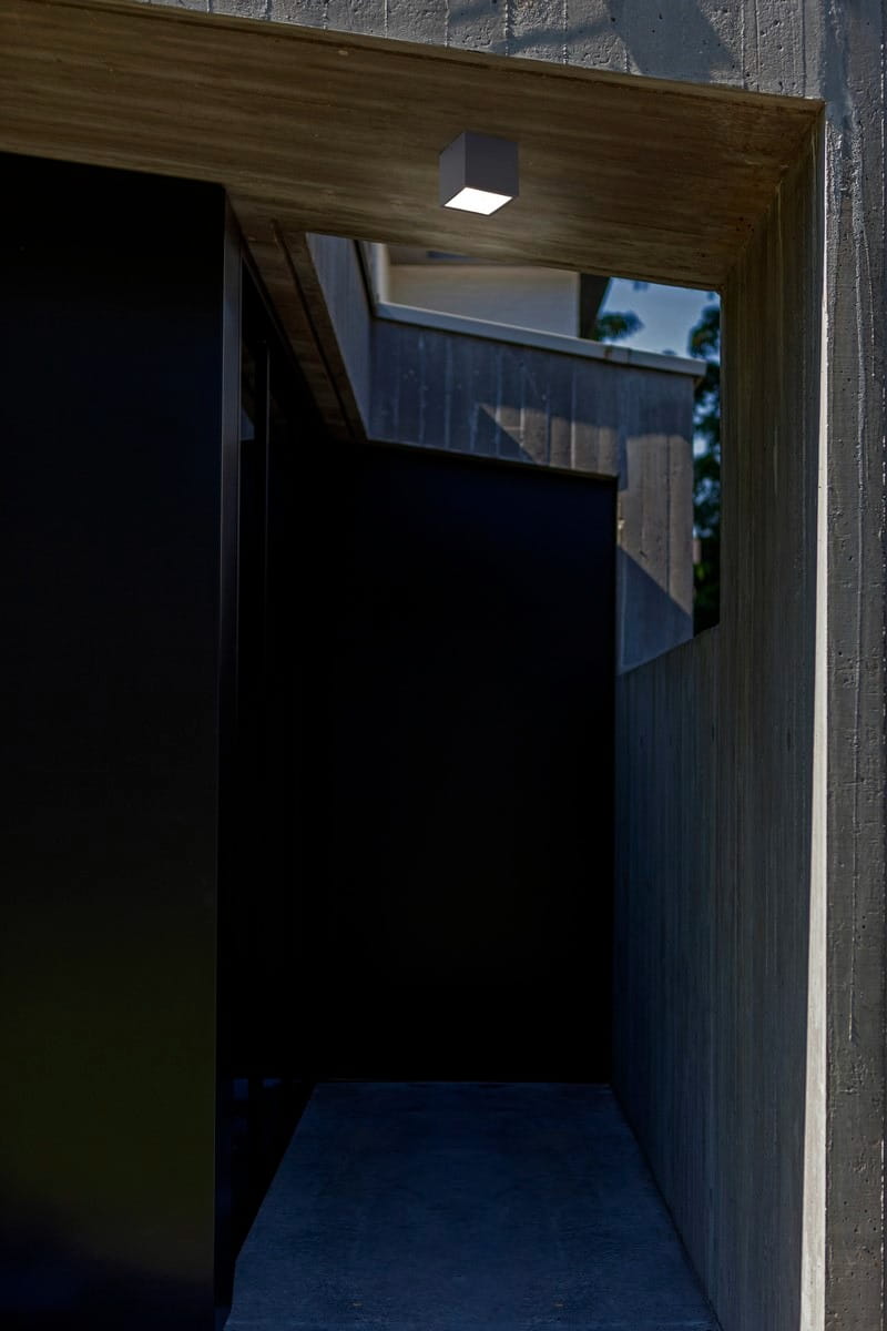 Ciemnoszara lampa sufitowa elewacyjna Gemini z jedną diodą LED 7W, 4000K. Produkt wiszący w prostokątnym kształcie, umieszczony w architektonicznym otoczeniu z betonu i czarnymi elementami. Jasne światło rozświetla wejście.