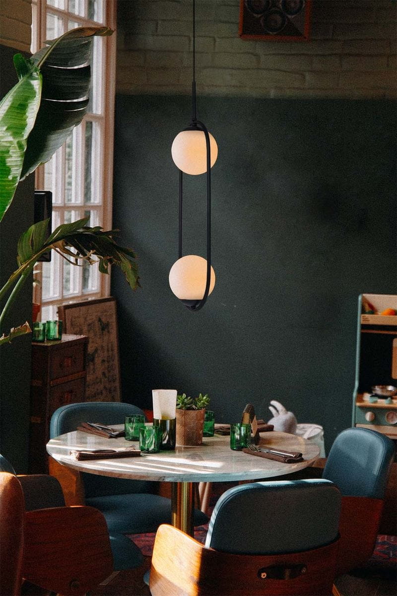 Lampa wisząca Parva 2xE14 w kolorze czarnym i białym, z dwoma okrągłymi kloszami, zawieszona nad stołem. W tle widać rośliny oraz meble w stylu retro, na stole zielone szklanki i dekoracyjna roślina.