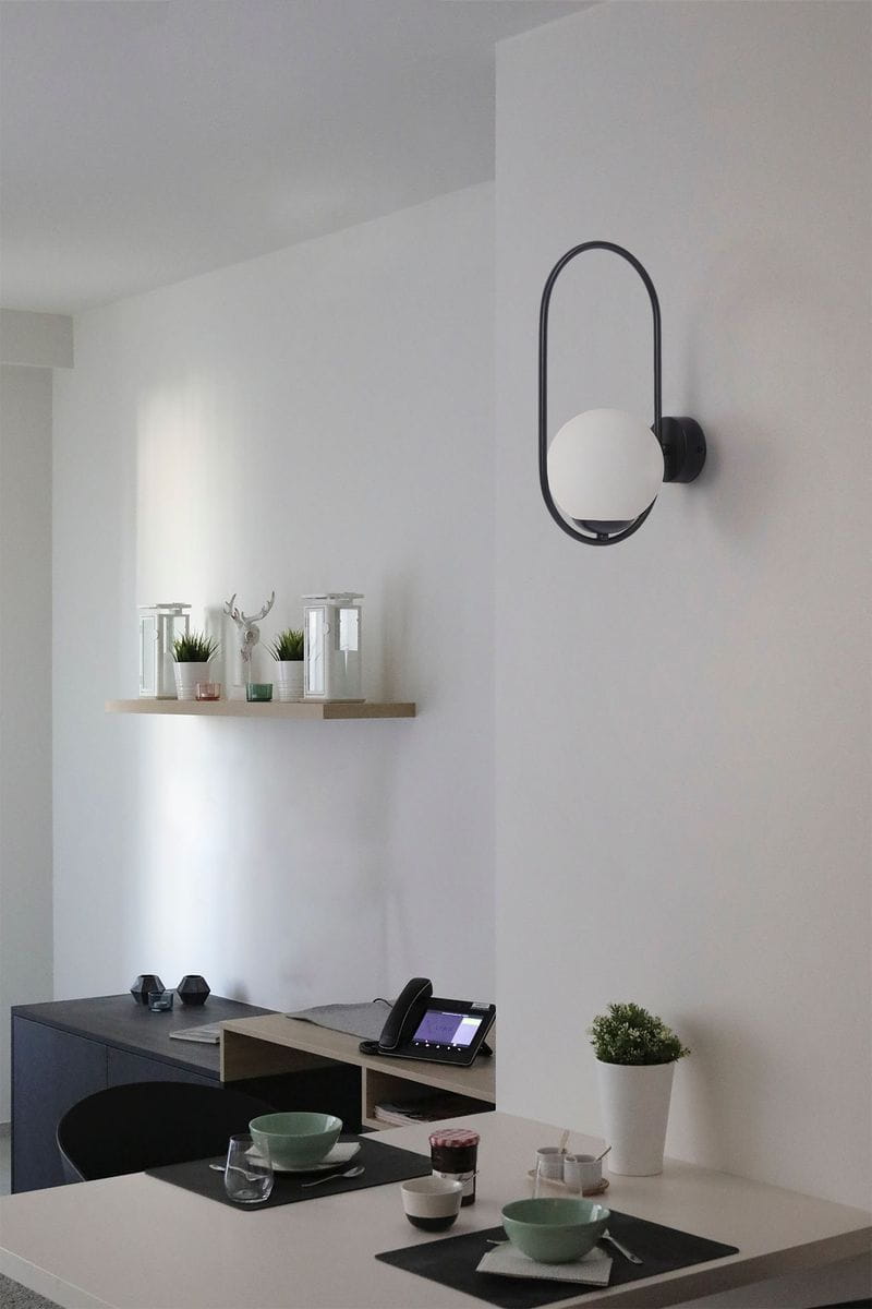 Kinkiet Parva 1xE14 w kolorze czarnym z białą, okrągłą kloszem. Montowany na ścianie, widoczny z boku. Otoczenie to nowoczesne wnętrze z drewnianą półką, czarnym stół i akcesoriami. Tło jasne, minimalistyczne.