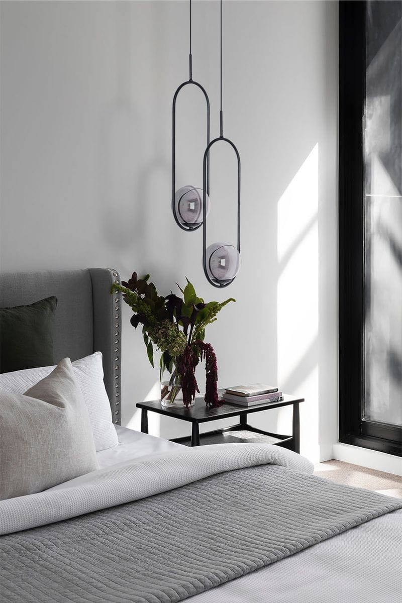 Lampa wisząca Davos 1xE14 w kolorze czarnym i grafitowym, o minimalistycznym kształcie, zawieszona w sypialni nad stolikiem nocnym. W tle widoczna dekoracyjna roślina oraz poduszki na łóżku, na jasnym tle.