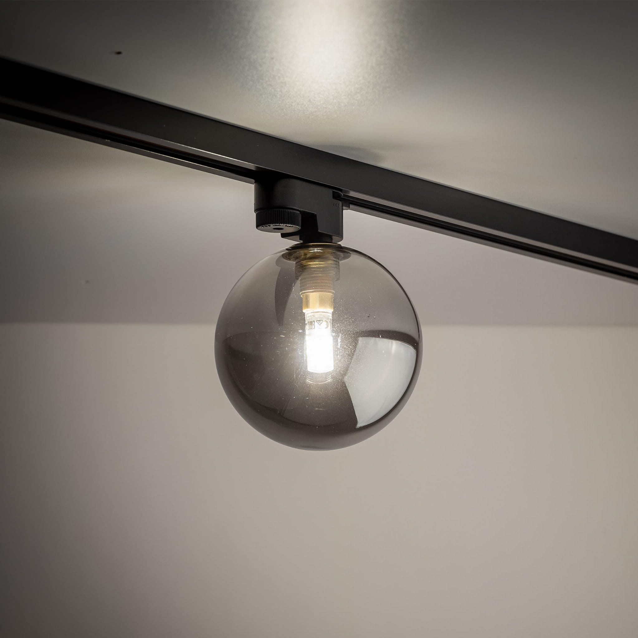 Lampa Tracer na szynę z jedną oprawą G9, w kolorze czarnym i grafitowym. Szklana kula z ciemnym szkłem, zawieszona na metalowej podstawie. Oświetlenie skierowane w dół, zamontowane na górnej krawędzi.
