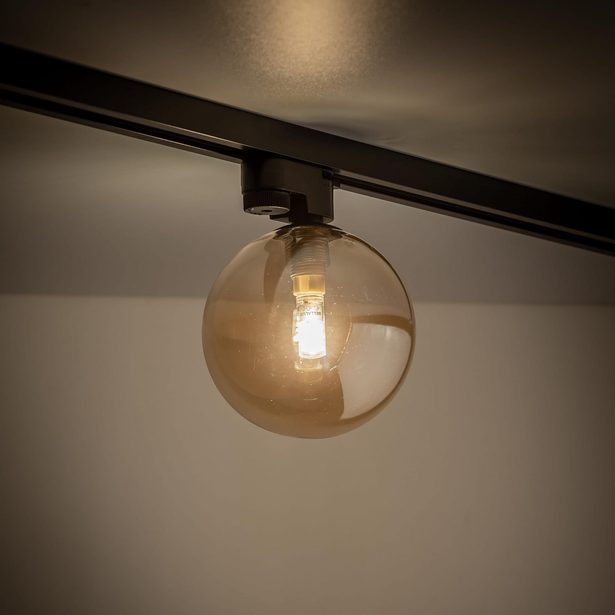 Lampa Tracer 1xG9 w kolorze czarnym z bursztynową, szklaną kulą. Widok z dołu, montowana na szynie, z widoczną żarówką. Tło w odcieniach szarości, podkreślające detale lampy.