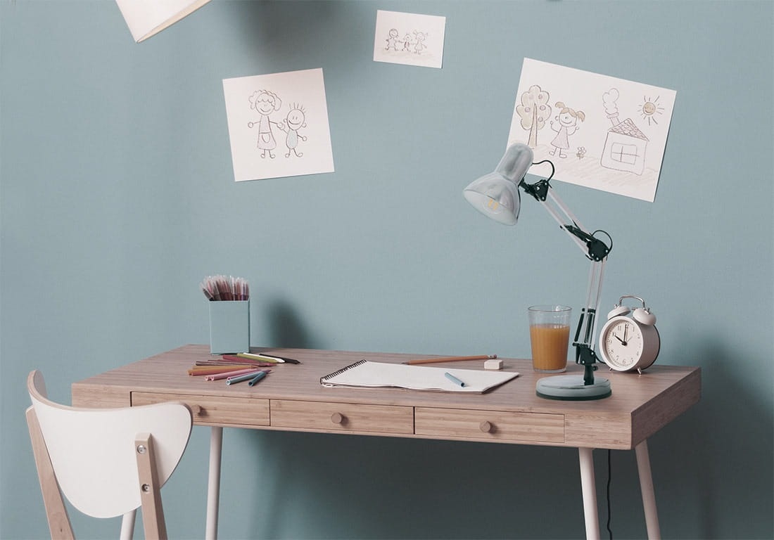 Lampka biurkowa Cosmo 1xE27 w kolorze chromu, z regulowanym ramieniem, stoi na drewnianym biurku. W tle błękitna ściana z rysunkami dzieci. Na biurku widoczne przybory do rysowania, szklanka z napojem i zegar.