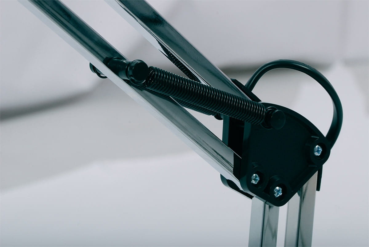 Lampka biurkowa Cosmo z chromowanym ramieniem, widok z boku. Widać metalowy mechanizm ruchomy oraz sprężynę dla regulacji wysokości. Tło proste, białe, podkreślające detale konstrukcji.