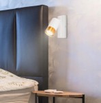 Klemens kinkiet 1xGU10 w kolorze białym i złotym, zamontowany na ścianie obok łóżka. Lampa skierowana w dół, z okrągłym kloszem, na tle ciemnej tapety. W pobliżu widoczna drewniana szafka z pilotem i książkami.