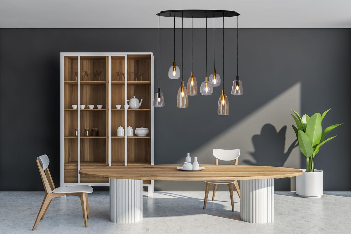 Lampa wisząca Divine z 8 żarówkami E14 w kolorze czarnym i multikolorowym, zawieszona na długich przewodach. Szklane klosze o różnych kształtach, umieszczone nad drewnianym stołem w nowoczesnym wnętrzu. Tło w odcieniach szarości.