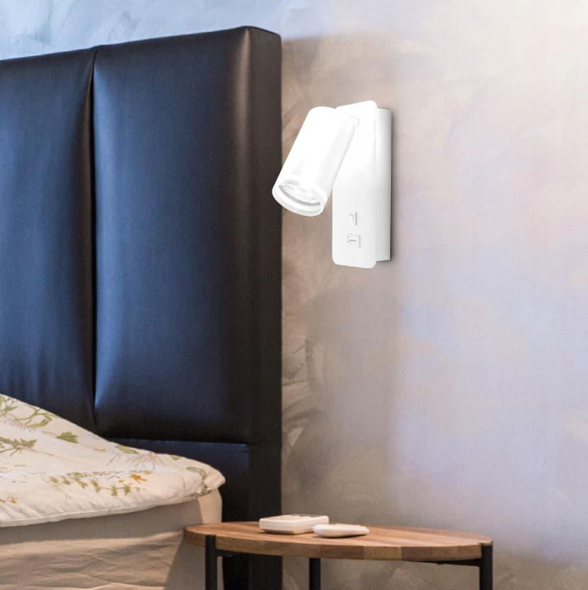 Kinkiet Ring z gniazdem USB, biały, montowany na ścianie, z ruchomą głowicą lampy GU10. Umieszczony obok łóżka na tle czarnej tapicerowanej ściany, z drewnianym stolikiem nocnym w dolnej części kadru.