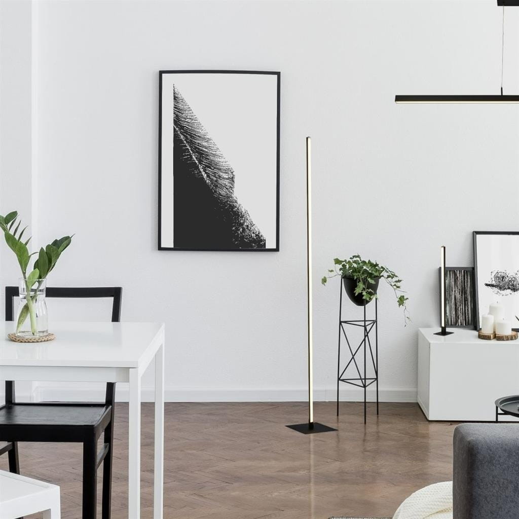 Lampa podłogowa Tribeca w kolorze czarnym, z jedną żarówką LED o mocy 18W. Smukła forma z prostokątną podstawą i cienkim korpusem, ustawiona w jasnym wnętrzu obok białego stolika i czarnego krzesła. W tle grafika na ścianie oraz roślina w doniczce.
