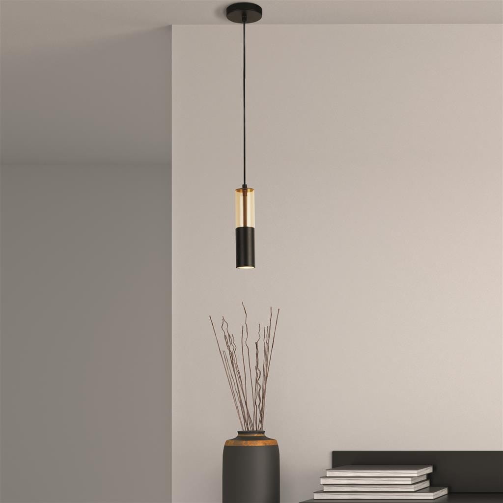 Lampa wisząca Merrygold 1xGU10 w kolorze czarnym i bursztynowym, zawieszona na cienkim kablu. Cylindryczny klosz z czarnej metalowej podstawy i bursztynowym szkle. W tle widoczny wazon z gałązkami oraz stół z książkami. Tło jasne, stonowane.
