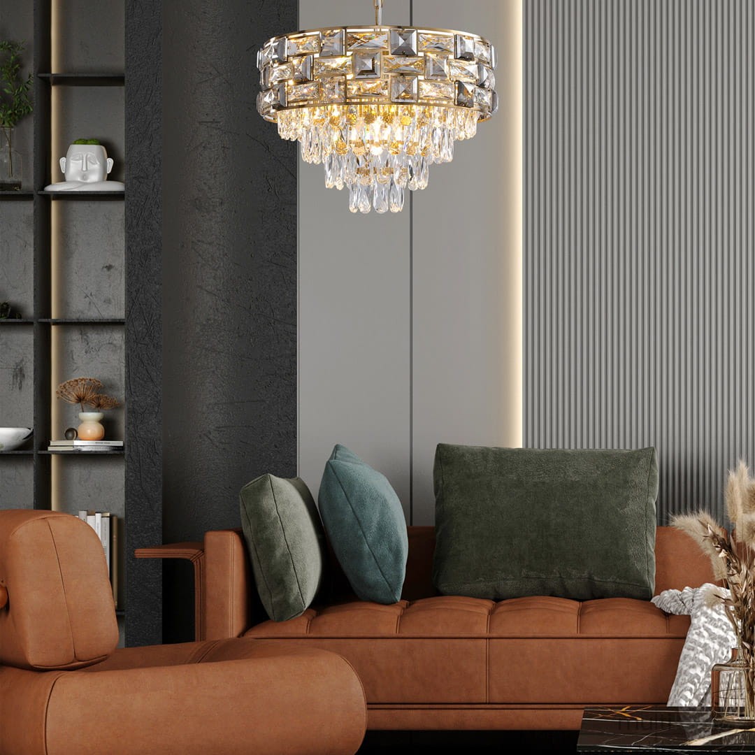 Lampa wisząca Luxuria 9xE14 w kolorze złotym, z kryształowymi elementami i ozdobnymi zwisami. Umieszczona w nowoczesnym wnętrzu nad brązową sofą z poduszkami w odcieniach zieleni. Ujęcie z boku, ukazujące detale konstrukcji.