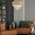 Lampa wisząca Luxuria 9xE14 w kolorze złotym, z kryształowymi elementami i ozdobnymi zwisami. Umieszczona w nowoczesnym wnętrzu nad brązową sofą z poduszkami w odcieniach zieleni. Ujęcie z boku, ukazujące detale konstrukcji.
