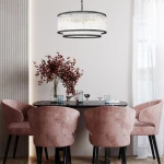Lampa wisząca Sergio 6xE14 w kolorze czarnym z przezroczystymi detalami, okrągła, zawieszona na łańcuchu. Umieszczona nad okrągłym czarnym stołem, w aranżacji z różowymi fotelami i dekoracyjnym bukietem. Tło jasne, elegancka atmosfera.