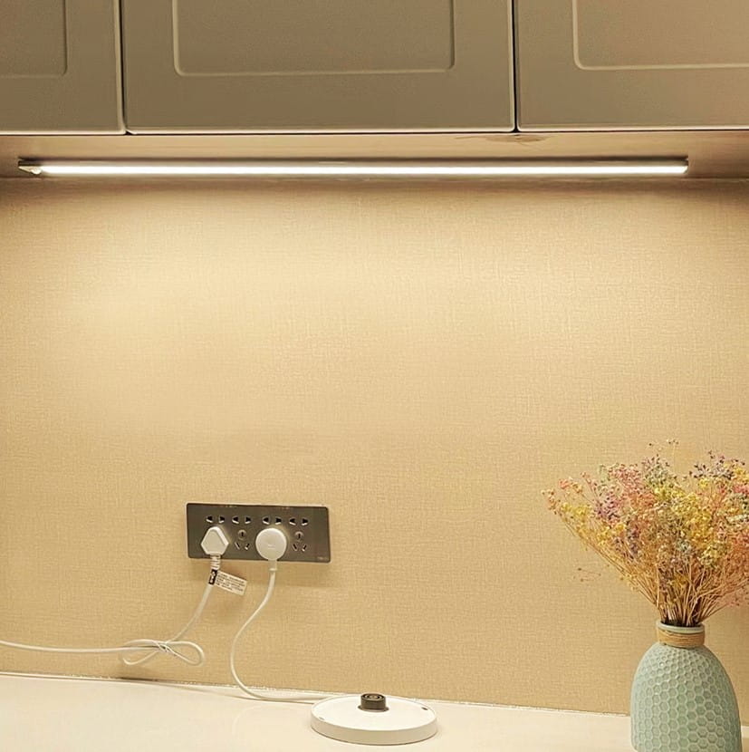 Listwa podszafkowa LED, czarna, 60 cm, z czujnikiem ruchu, zamontowana nad blatem. Widoczna włączona lampa, gniazdko elektryczne oraz dekoracyjny wazon z kwiatami na białym stole. Tło w jasnym kolorze.