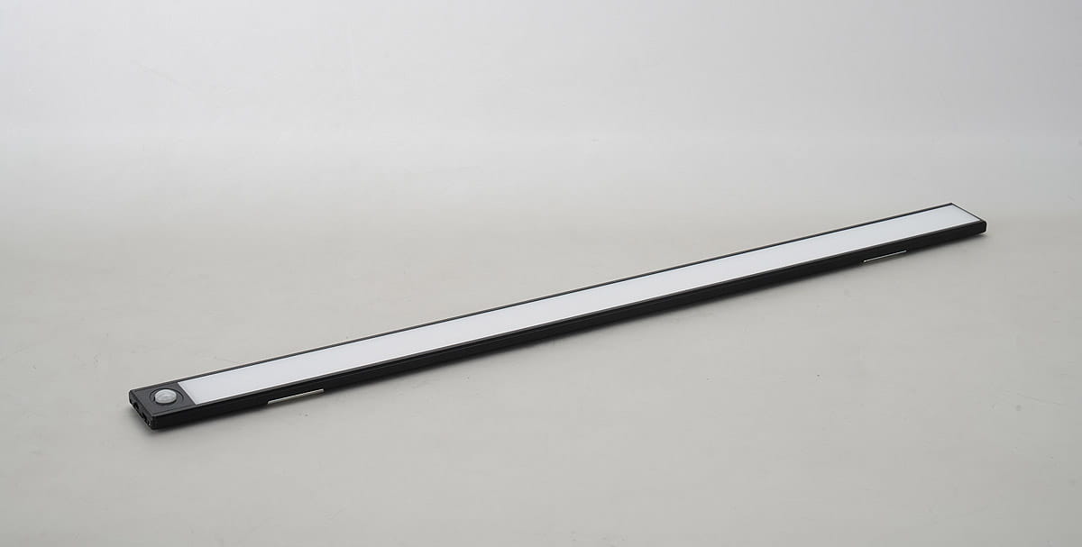 Listwa podszafkowa LED 60 cm w kolorze czarnym z wbudowanym czujnikiem ruchu. Produkt leży na szarej powierzchni, widoczny z boku, z białym panelem świetlnym i przyciskiem z lewej strony. Służy do oświetlenia przestrzeni użytkowej.
