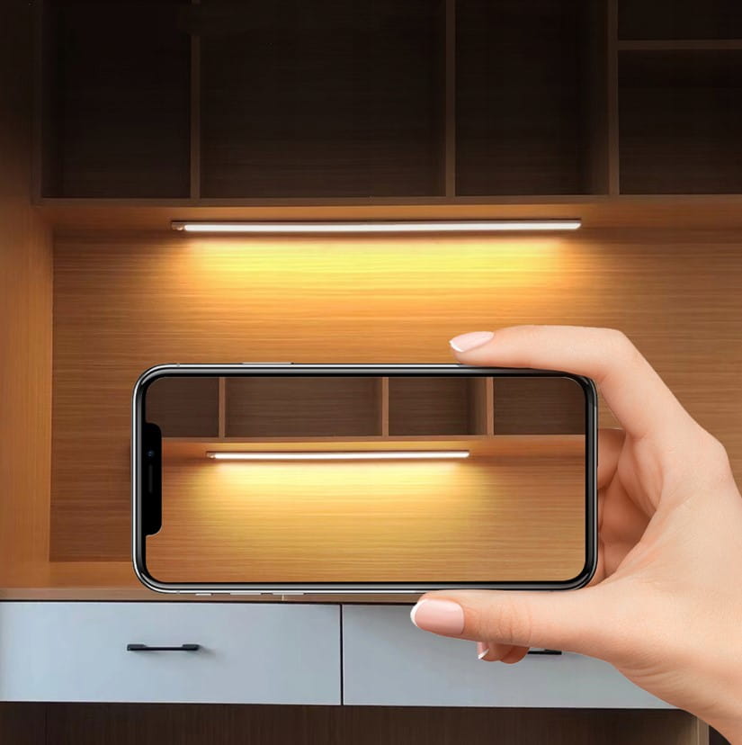 Srebrna listwa podszafkowa LED o długości 80 cm z czujnikiem ruchu, zamontowana pod półką, oświetlająca przestrzeń. Obok trzymany telefon, na tle drewnianej ściany z szafkami.