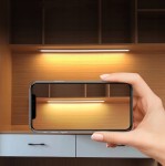 Srebrna listwa podszafkowa LED o długości 80 cm z czujnikiem ruchu, zamontowana pod półką, oświetlająca przestrzeń. Obok trzymany telefon, na tle drewnianej ściany z szafkami.