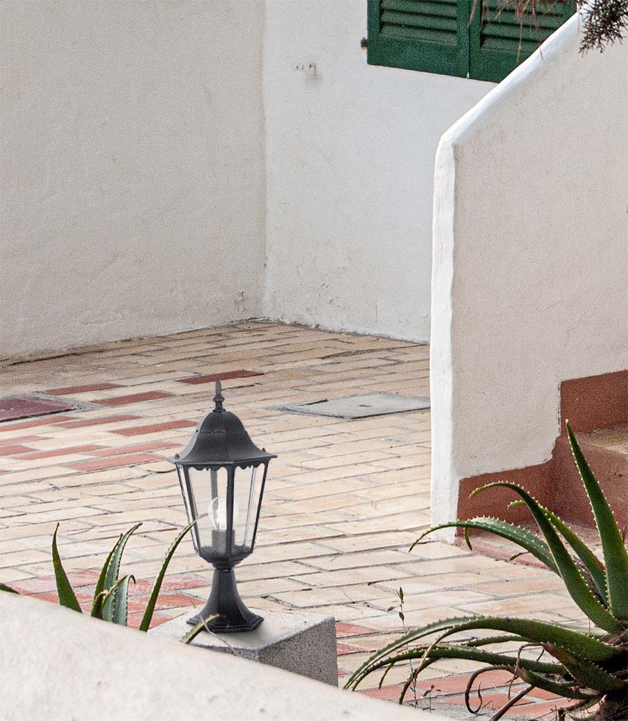 Czarna lampa ogrodowa Lozana H51 stojąca, wykonana z metalu, z klasycznym, sześciokątnym kloszem. Ujęcie z boku, na płaskim podłożu, w otoczeniu roślin. Tło jasne, z widocznymi elementami architektonicznymi.
