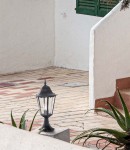 Czarna lampa ogrodowa Lozana H51 stojąca, wykonana z metalu, z klasycznym, sześciokątnym kloszem. Ujęcie z boku, na płaskim podłożu, w otoczeniu roślin. Tło jasne, z widocznymi elementami architektonicznymi.
