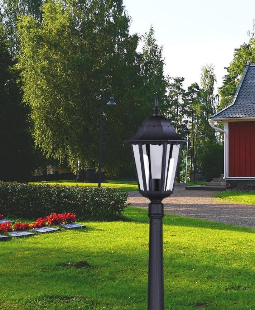 Lampa ogrodowa stojąca Lozana H114 w kolorze czarnym, wykonana z metalu. Posiada klasyczny kształt z przezroczystymi panelami, umieszczona na wysokim słupku, otoczona zielonym trawnikiem i kwiatami w tle.