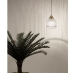 Lampa wisząca Ahigal 1xE14 w kolorze złotym z przezroczystym kloszem o żłobionej fakturze. Wisi na cienkim kablu, w tle widoczna roślina doniczkowa, a ściana za lampą ma delikatną, teksturowaną powierzchnię.