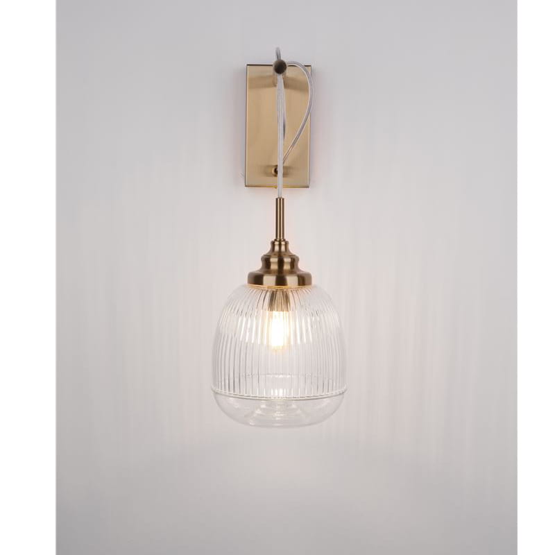 Kinkiet Ahigal 1xE14 w złotej oprawie, z przezroczystym kloszem o teksturowanej powierzchni. Zawieszony na prostokątnej, metalowej podstawie. Źródło światła widoczne wewnątrz klosza, umieszczony na białym tle.