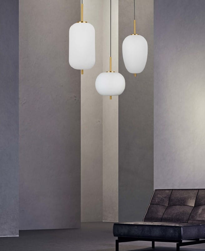 Lampa wisząca Pilar 3xE14 w kolorze złotym, składająca się z trzech mlecznych kloszy w różnych kształtach. Ujęcie frontalne, zawieszona na cienkich linkach, w minimalistycznym wnętrzu z szarymi ścianami i czarną sofą.