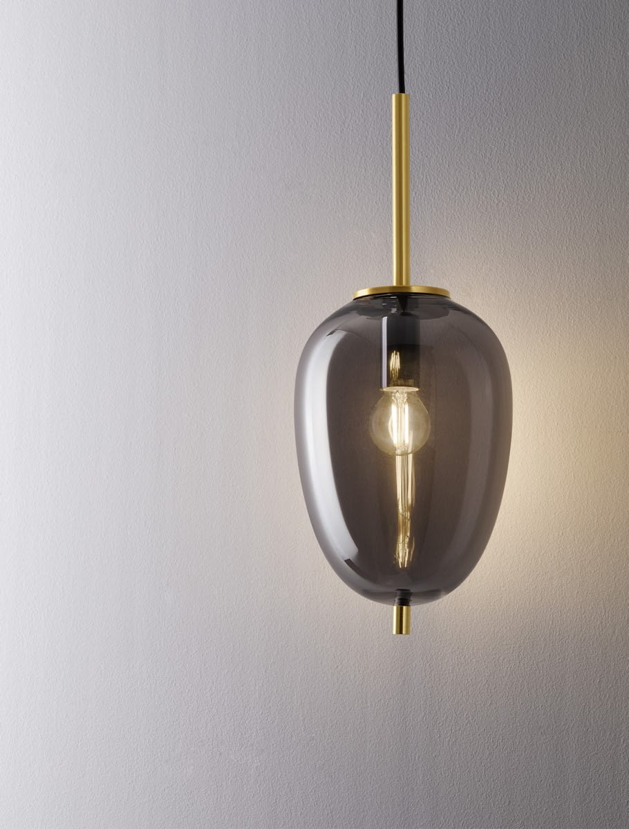 Lampa wisząca Pilar L o średnicy 15,8 cm, z przezroczystym kloszem w odcieniu szaro-czarnym i złotymi detalami. Umieszczona na cienkim, czarnym przewodzie, z widoczną żarówką E14 wewnątrz. Produkt zawieszony w pozycji pionowej, na jasnym tle.